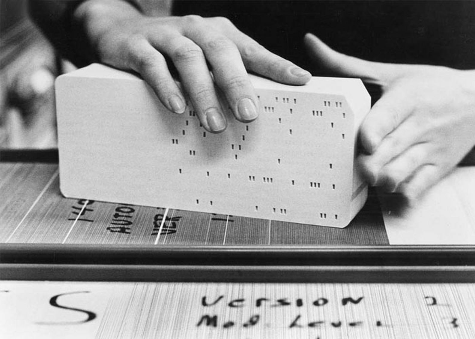 Hands handling a punched card on a table, with version notes written by hand